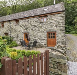 An outdoor patio area with table and chairs in front of a stone cottage with wooden doors and windows at Hendoll Cottage 2 Fairbourne near Dolgellau