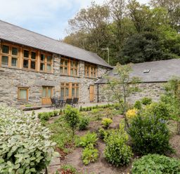 A stone house with wooden windows and doors next to a garden with plants and shrubs at Hendoll Barn Friog near Fairbourne