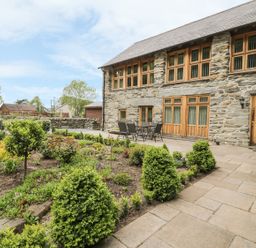 A stone house with wooden windows and doors next to a garden and patio with table and chairs at Hendoll Barn Friog near Fairbourne