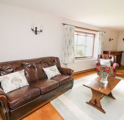 A living room with a leather sofa and armchair a wooden coffee table with flowers and floral curtains at Holly Farm Cottage in Howey