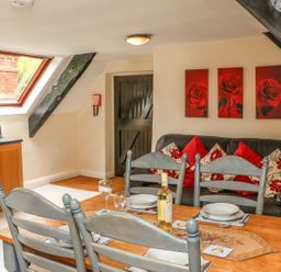 A dining area with a wooden table and chairs set for four with a bottle of wine and a black leather sofa with red and floral cushions in a room with a skylight window and wooden cabinets at Fig Trees - Wibble Farm in Williton near Watchet