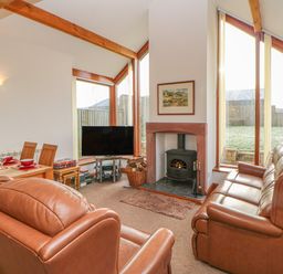A living room with leather sofas a wood-burning stove a television wooden dining table and large windows at Swallow Cottage Milton near Brampton