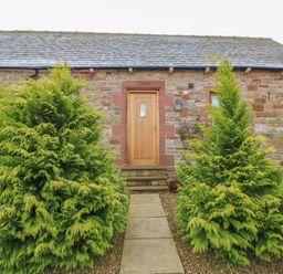 The front entrance of a stone cottage with a wooden door and two large green shrubs on either side of a stone path at Swallow Cottage in Milton near Brampton