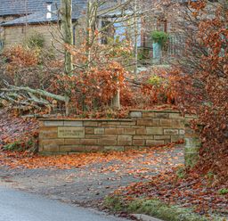 A stone wall with a sign saying Milton Mains Cottages surrounded by trees with autumn leaves on the ground at Swallow Cottage in Milton near Brampton