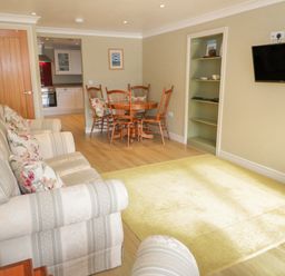 A living room with patterned sofas a wooden dining table with chairs a wall-mounted TV and a kitchen visible through a doorway at Bryn Y Don Cottage in Benllech