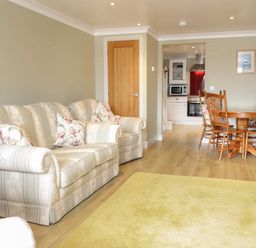 A living room with a striped sofa, floral cushions, wooden dining table with chairs and kitchen in the background at Bryn Y Don Cottage in Benllech