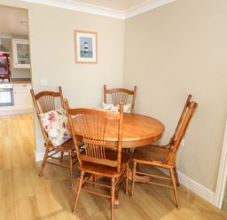 A dining area with a round wooden table and four wooden chairs with cushions next to a kitchen with white cabinets at Bryn Y Don Cottage in Benllech