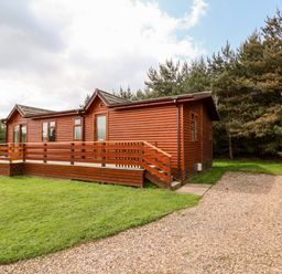 A wooden lodge with a fenced porch next to a gravel driveway and surrounded by grass and trees at Callow Lodge 5 in Beaconsfield Holiday Park