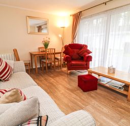 A living room with a sofa and armchair near a wooden table with chairs and a large window with curtains at Peacehaven Rosecraddoc Lodge near Liskeard