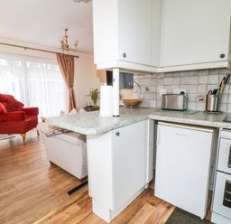 A kitchen counter with white cabinets and appliances next to a living area with a red armchair and wooden floor at Peacehaven Rosecraddoc Lodge near Liskeard