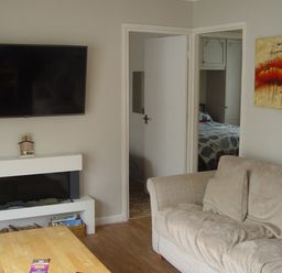 A living room with a mounted TV above a fireplace a beige couch with cushions a wooden coffee table and an open door showing a bedroom at Peacehaven Rosecraddoc Lodge near Liskeard