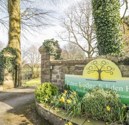 Entrance gate with stone pillars covered in ivy and a sign for Eden Lodge and Eden House surrounded by plants and daffodils at Eden Lodge in Bardsea