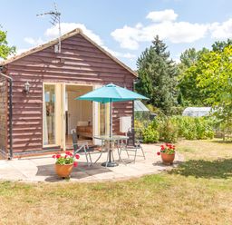 A small wooden house with a patio table, chairs, and umbrella in a garden at Norbank Garden Studio in Bressingham