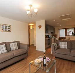 A living room with two brown sofas a glass coffee table and an open kitchen at Roan Cottage in Royal Wootton Bassett