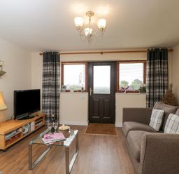 A living room with a brown sofa and checkered pillows a glass coffee table wooden tv stand with a lamp a television and wall art at Roan Cottage in Royal Wootton Bassett
