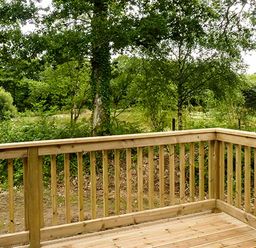 A wooden deck railing overlooking a forested area with trees and bushes at Chaffinch Lodge in Highampton near Sheepwash