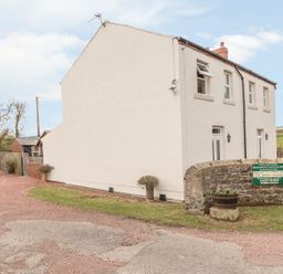 An exterior view of a two-story white cottage with windows and doors a stone wall with a sign and a planter in front at St Cuthberts Cottage Beal near Holy Island