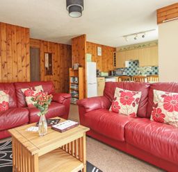 A living room with red leather sofas and floral cushions a wooden coffee table and a kitchen area at 4 Manorcombe in Honicombe Manor near Gunnislake