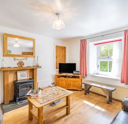 A living room with a wooden coffee table and puzzle pieces a fireplace with a large mirror and two bottles a television on a wooden stand a window with red checked curtains and a wooden bench at No 7 Spark Bridge near Ulverston