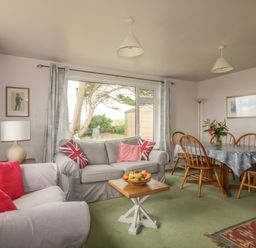 A living room with sofas, a wooden dining table with chairs, a lamp, and a window with curtains at Quarterdeck in Bigbury-on-Sea near Modbury