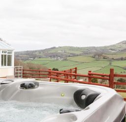 An outdoor hot tub with bubbles next to a conservatory and a wooden fence overlooking green hills at Bryn Derw Holiday Cottage in Eglwysbach near Llanrwst and Conwy