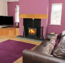 A living room with a brown leather sofa, purple cushions, a fireplace with fire, two windows, a purple rug, and a television on a wooden cabinet at Bryn Derw Holiday Cottage in Eglwysbach near Llanrwst and Conwy