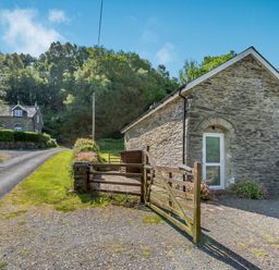 A stone building with a wooden gate and gravel driveway near a road and trees at Cwmdu Cottage in Cwm-Cou near Newcastle Emlyn
