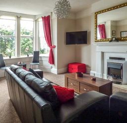 A living room with a black leather sofa red curtains a wooden coffee table a wall-mounted TV and a fireplace at Westgate Retreat in Hunstanton