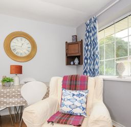 A dining area with a polka dot tablecloth on a table surrounded by white chairs and a cream armchair with a fish-pattern pillow near a window with blue and white curtains at No 1 The Heathers Rosecraddoc Lodge near Liskeard