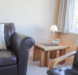 A corner of a living room with a leather sofa and armchair a wooden side table with a lamp a picture frame and a decorative love sign near a window at Tegfan in Trearddur Bay