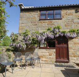 An outdoor patio with four chairs and a wooden picnic table next to a stone building with a wooden door and windows with flowering plants above at Goathland Cottage in Ruswarp near Whitby