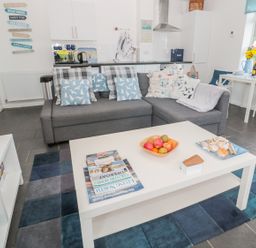 A living room with a gray sofa and blue patterned cushions a white coffee table with magazines fruit and seashells a blue rug and a white dining table with chairs by a window at Bracken Barn Cottage in Widdrington near Amble