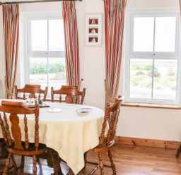 A dining room with a wooden table and chairs near two windows with striped curtains at McGuire's Cottage in Barnatra County Mayo
