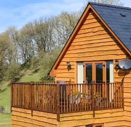 A wooden cabin with a balcony and railing in a grassy area with trees at Brynhir Farm in Howey near Llandrindod Wells