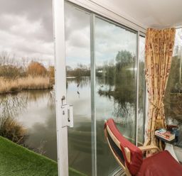 A red chair and small table by a large glass door overlooking a lake with reeds and trees at Secret Island Yurt in Beckford near Tewkesbury