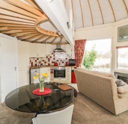 An open plan kitchen and living area with a round glass dining table a beige sofa and a fireplace at Secret Island Yurt Beckford near Tewkesbury