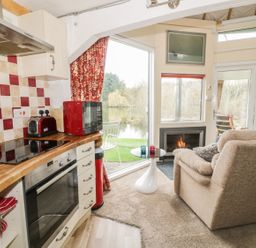 A kitchen with a wooden countertop, red kettle, toaster, microwave, and oven next to a living area with a beige sofa, small round white table, fireplace, and glass doors showing an outside view at Secret Island Yurt in Beckford near Tewkesbury