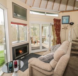 A living area with a beige sofa near a fireplace and glass doors with views of a river at Secret Island Yurt in Beckford near Tewkesbury