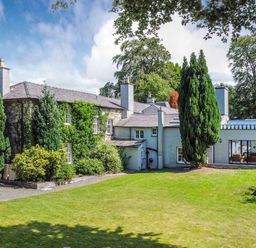 An exterior view of a large stone and white two-story house with multiple chimneys surrounded by tall trees and a lawn at Bryn Derwen Bethesda near Caernarfon