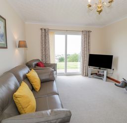 A living room with leather sofas and yellow cushions a television on a stand and glass sliding doors at Bryn Coch Bach in Dyffryn Ardudwy