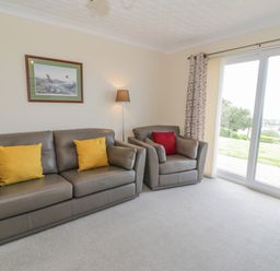 A living room with two leather sofas and cushions a floor lamp a painting on the wall and sliding glass doors with curtains at Bryn Coch Bach in Dyffryn Ardudwy
