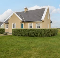 A beige house with gray roof and trimmed hedges in front on a grassy lawn at 8 Lios Na Sioga in Binghamstown near Belmullet County Mayo