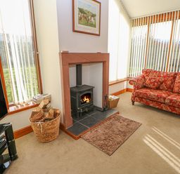 A living room with a wood burning stove a basket of firewood a red patterned sofa and windows with vertical blinds at Swift Cottage in Milton near Brampton