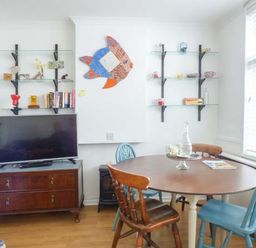 A dining area with a round wooden table and mixed wooden chairs next to a TV on a wooden cabinet and wall shelves at Tidewood in Polzeath