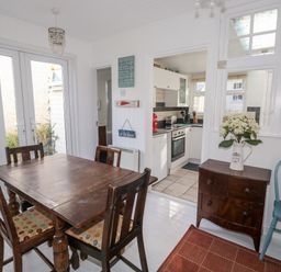 A dining area with a wooden table and four chairs next to a kitchen with white cabinets and appliances at Driftwood Polzeath near Wadebridge