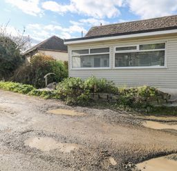 A gravel road with puddles beside a building with horizontal siding and large windows at Driftwood in Polzeath near Wadebridge