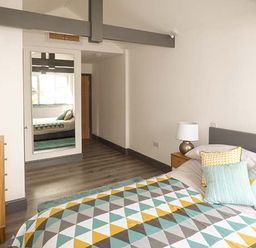 A bedroom with a bed covered in a geometric pattern bedspread and pillows a wooden chest of drawers with a blue vase a bedside table with a lamp and a mirror on the wall at White Cottage Annexe in Tenby