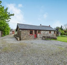 A stone cottage with a slate roof and red door surrounded by gravel driveway and green trees at Cilwen Fach in Cynwyl Elfed