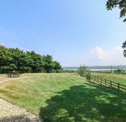 A grassy backyard with a wooden picnic table and trees near a fence overlooking a lake at Fferam Y Llyn in Rhosgoch near Cemaes Bay