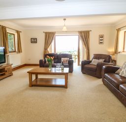 A living room with brown leather sofas a wooden coffee table with flowers and glasses a wooden TV stand with a television and windows with curtains at Fferam Y Llyn in Rhosgoch near Cemaes Bay